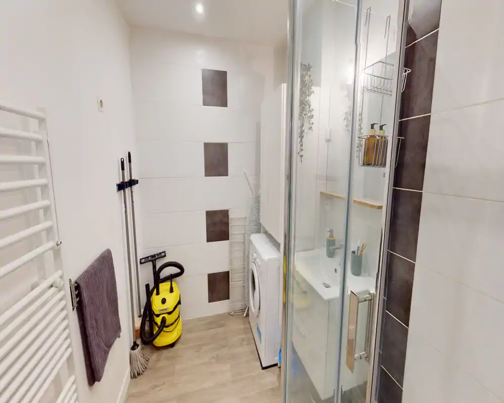 Modern bathroom with glass shower enclosure, vanity sink and a washing machine; white tiles and wood-effect flooring create a clean, functional space.