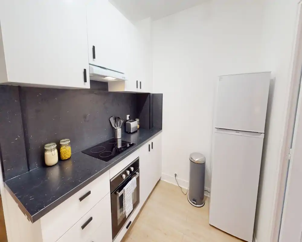 Modern compact kitchen with white cabinets, black marble-effect countertop and integrated electric hob and oven. Bright, clean space with small appliances and pantry jars on the counter.