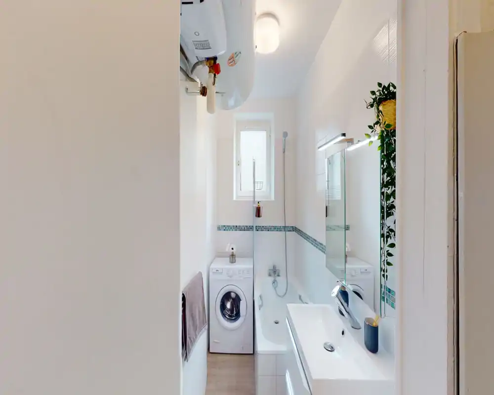 Compact, modern bathroom with a bathtub, wall-mounted shower, sink with mirror, and a washing machine. Clean white tiles, decorative mosaic border and a hanging plant add a fresh, tidy look.