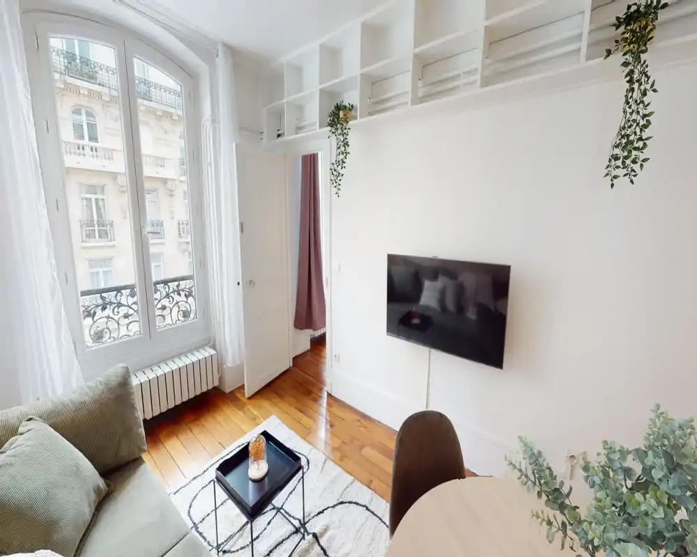 Bright, stylish living room with a comfortable sofa, rug and a wall-mounted TV. Large arched window fills the space with natural light and elegant Parisian details are visible outside.
