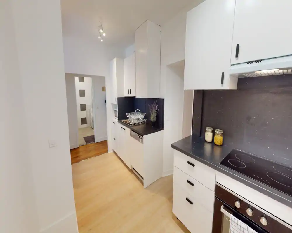 Modern, compact kitchen with white cabinets, black countertops and integrated appliances. Bright, functional layout with a ceramic hob, oven and neat storage including jars and a dish rack.