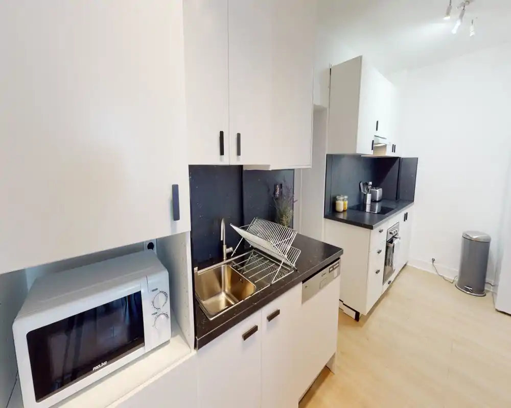 Modern compact kitchen with white cabinetry, black countertops, stainless sink and built-in appliances; bright, clean and efficiently arranged for everyday use.