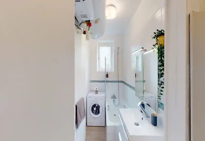 Compact, modern bathroom with a bathtub, wall-mounted shower, sink with mirror, and a washing machine. Clean white tiles, decorative mosaic border and a hanging plant add a fresh, tidy look.