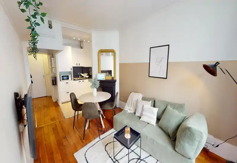 Bright, stylish living area with a comfortable green sofa, round dining table and an open kitchenette — warm wooden floors and tasteful decor create a welcoming atmosphere.
