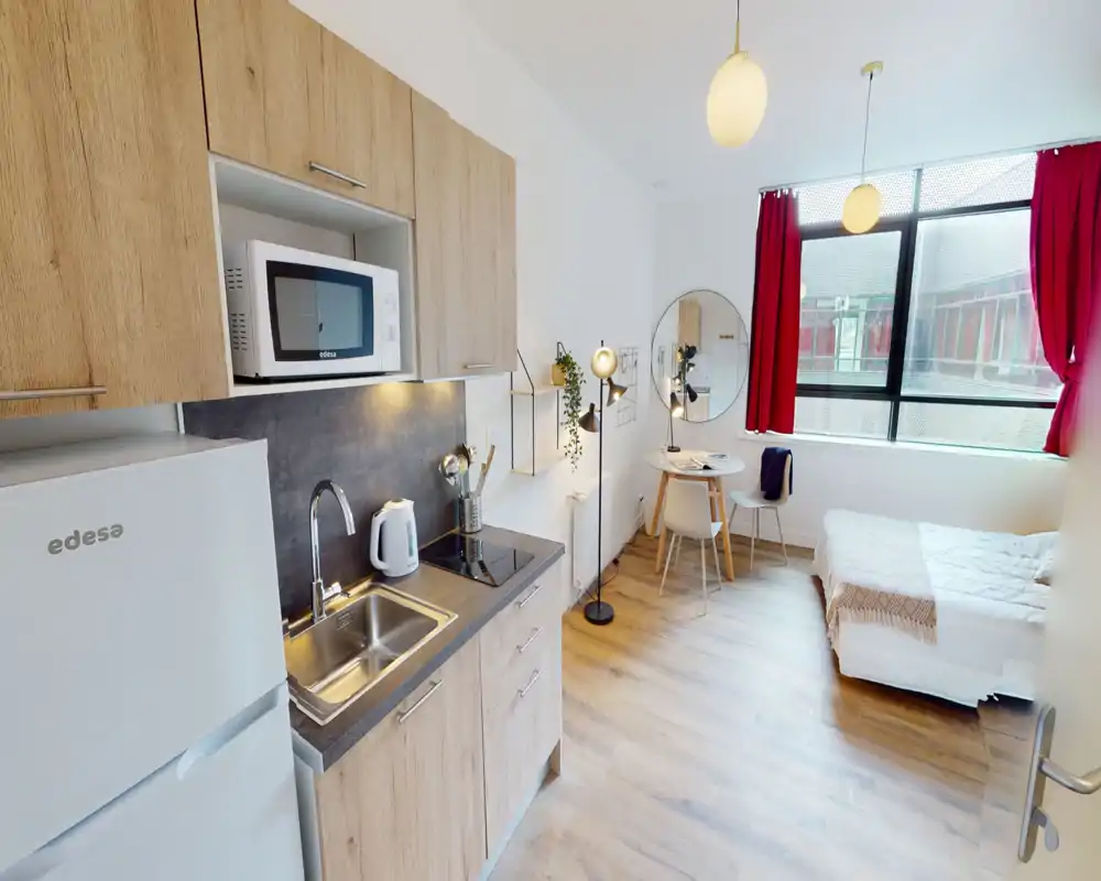 Studio lumineux et moderne avec coin cuisine intégré. Une grande fenêtre inonde l'espace de lumière ; lit, coin repas et kitchenette sont visibles dans une disposition soignée.