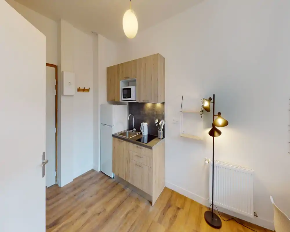 Kitchenette compacte avec placards effet bois, évier, plaque à induction, micro-ondes et réfrigérateur. Le sol en bois et l'éclairage doux créent une atmosphère chaleureuse et soignée, parfaite pour des locations courte durée.