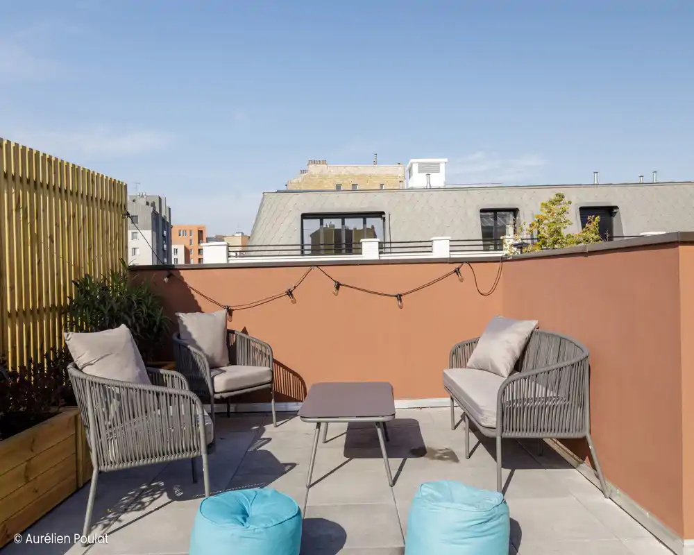 Terrasse extérieure avec sièges et un bas mur-rideau ; le ciel et les toits voisins sont visibles. Remarque : la photo présente des artefacts numériques mais montre un coin extérieur avec des sièges.