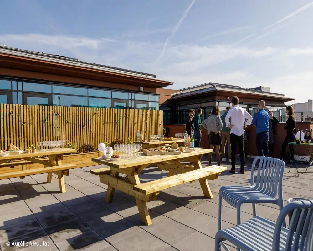 Terrasse extérieure ensoleillée avec longues tables de pique-nique en bois et bancs, places pour groupes et ciel dégagé — idéale pour repas collectifs et événements.
