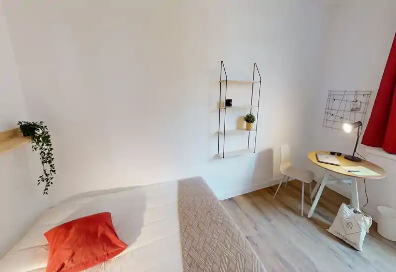Bright, minimalist bedroom with a neatly made bed, red accent pillow and a small workspace by the window. Clean white walls, wooden floor and decorative shelving create a calm, cosy atmosphere.