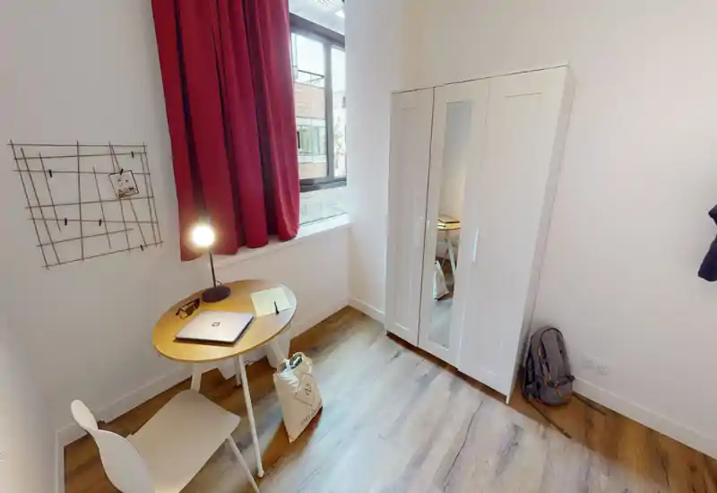 Compact bedroom corner with a small desk and wardrobe. The room features wooden flooring, a red curtain over a bright window, a white three-door wardrobe with a mirrored panel, and a minimalist round table with a lamp—ideal for a single occupant.