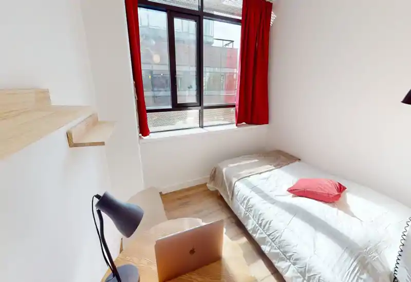 Bright single bedroom with a comfortable bed beside a large window with red curtains; wooden floor and minimal desk create a neat, airy feel ideal for short-term rentals.