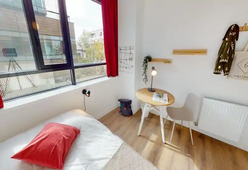 Bright, modern bedroom corner with a neatly made bed, red accent pillow and large window providing abundant natural light; a small round table and chair create a cozy workspace.