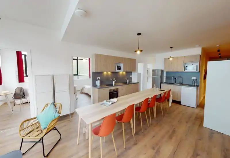 Spacious modern kitchen with a long wooden island and multiple orange dining chairs, integrated appliances and warm wood cabinetry — bright, clean and great for group dining.
