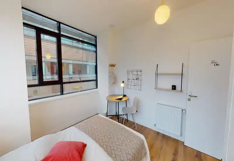 Dormitorio individual moderno y luminoso con la cama bien hecha, una pequeña mesa redonda junto a la ventana y estanterías minimalistas — ideal para estudiantes o profesionales.