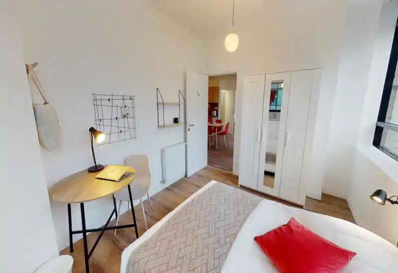 Dormitorio moderno y luminoso con cama doble vestida en blanco y plaid decorativo, lámpara, pequeña mesa de trabajo con silla y armario con espejo. Ventana amplia que aporta luz natural y suelo de madera que aporta calidez — ideal para alquileres de corta estancia.