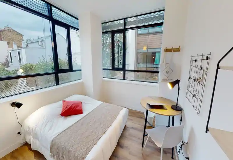 Dormitorio moderno y luminoso con grandes ventanas en esquina, cama doble impecable con almohadón rojo y una pequeña mesa de trabajo — perfecto para descansar y teletrabajar.