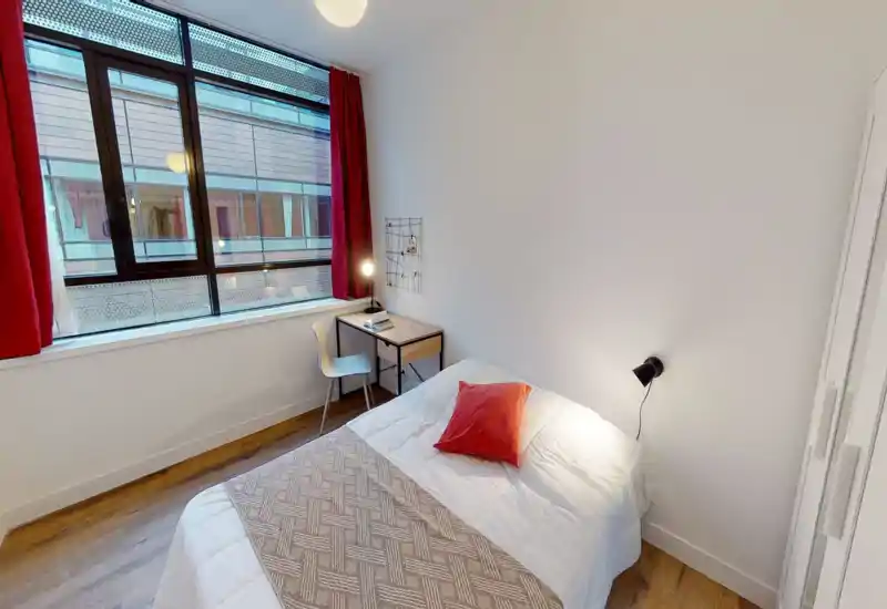 Bright, neatly staged single bedroom with a comfortable bed, red accent cushion and a small desk by a large window that fills the room with natural light.