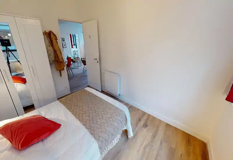 Bright, simply styled bedroom with a visible double bed, textured throw and a red accent cushion. Light wood floor and a wardrobe with mirrored doors are shown; door opens onto a living area.
