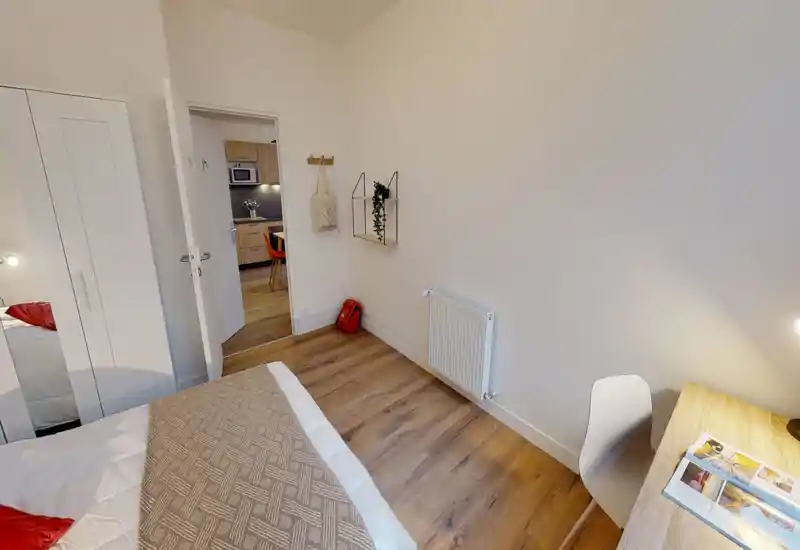 Camera da letto luminosa e semplice, con ampio letto in primo piano, pavimento in legno e arredi moderni essenziali tra cui scrivania e armadio. Illuminazione calda e pareti neutre creano un ambiente tranquillo, ideale per un annuncio.