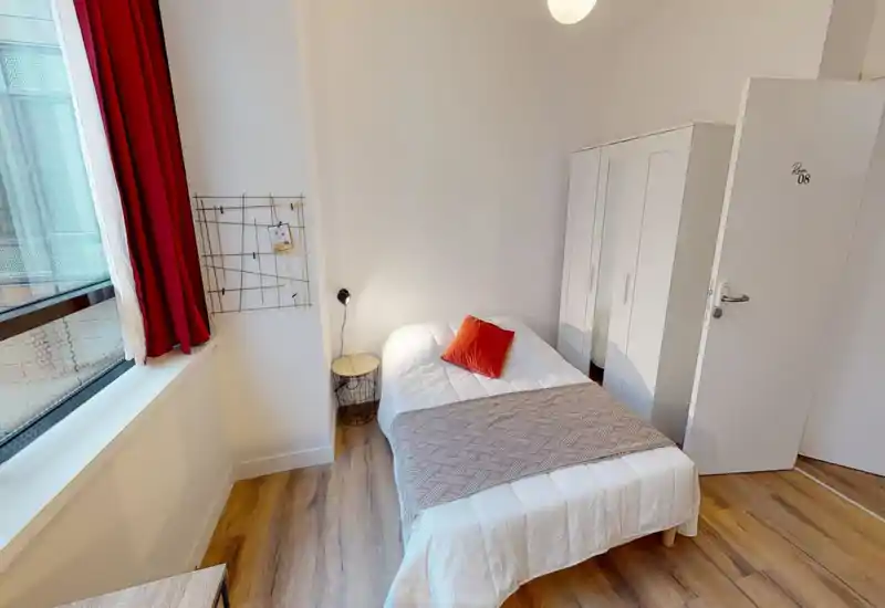 Camera da letto luminosa e ordinata con un letto matrimoniale con biancheria bianca, un plaid e un cuscino rosso. La stanza include un armadio, un comodino con lampada e una grande finestra che garantisce luce naturale.