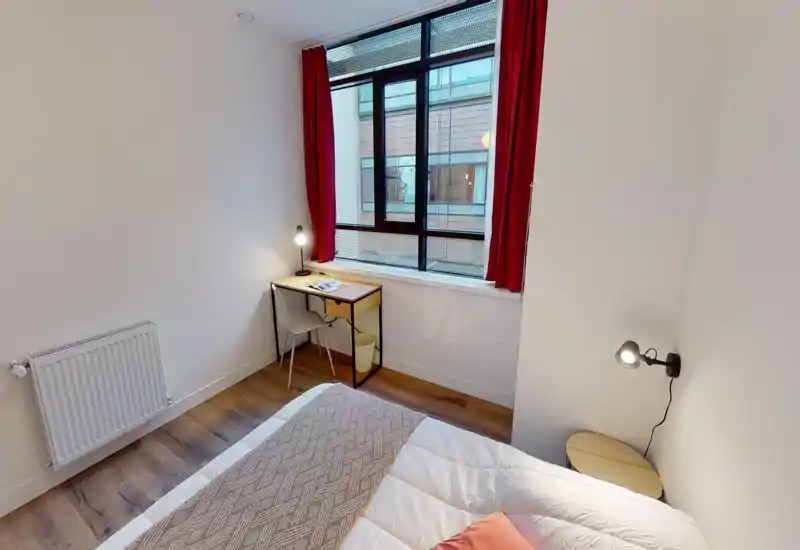 Camera da letto luminosa e arredata in modo semplice con letto visibile, lampada da comodino e una piccola scrivania vicino alla finestra con tende rosse. Stile pulito e moderno, buona luce naturale, adatta per annunci di affitto.
