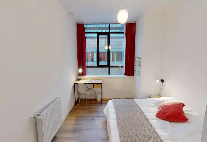 Camera da letto luminosa e ben arredata con letto matrimoniale, lampada da comodino e una piccola scrivania vicino alla finestra con tende rosse — perfetta per una persona o una coppia che cerca una stanza moderna e compatta.