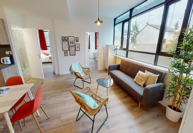 Spacious, sunlit living room with a modern grey sofa, rattan chairs, a small coffee table and large windows that flood the space with natural light — stylish and inviting for relaxing or entertaining.