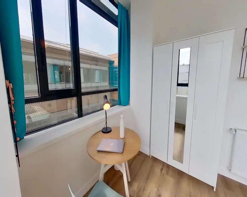 Bright corner living area with a round wooden table, lamp and wardrobe by a large window with turquoise curtains, ideal for a small lounge or study nook.