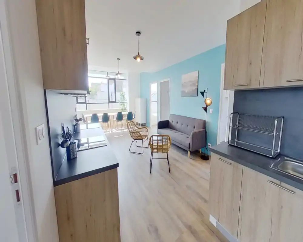 Bright open-plan living area with a small modern kitchen in the foreground, a grey sofa, rattan chairs and a dining table by the window; ideal for social living and natural light.