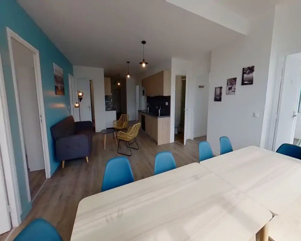 Spacious open-plan living area with a dining table in the foreground, a lounge with a sofa and rattan chairs, and a compact kitchen along the back wall — modern, bright and well coordinated.