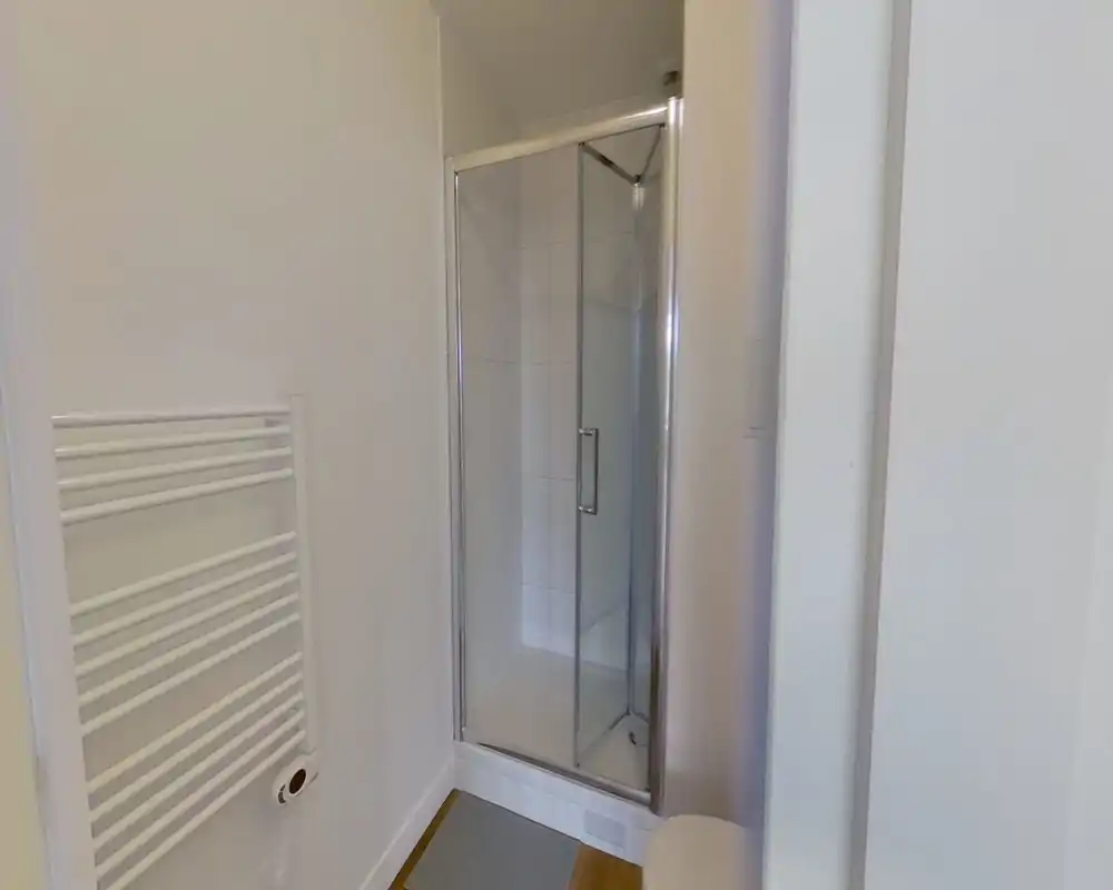 Compact bathroom with a tiled corner shower cubicle, glass door and a wall-mounted heated towel rail. Bright neutral finishes and simple, modern fittings.