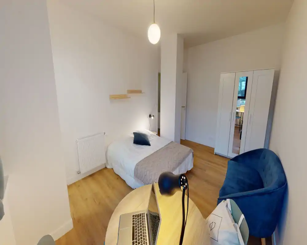 Dormitorio luminoso y sencillo con cama doble, suelo de madera, un pequeño sofá azul y armario con espejo; ideal para alquiler individual o pareja.