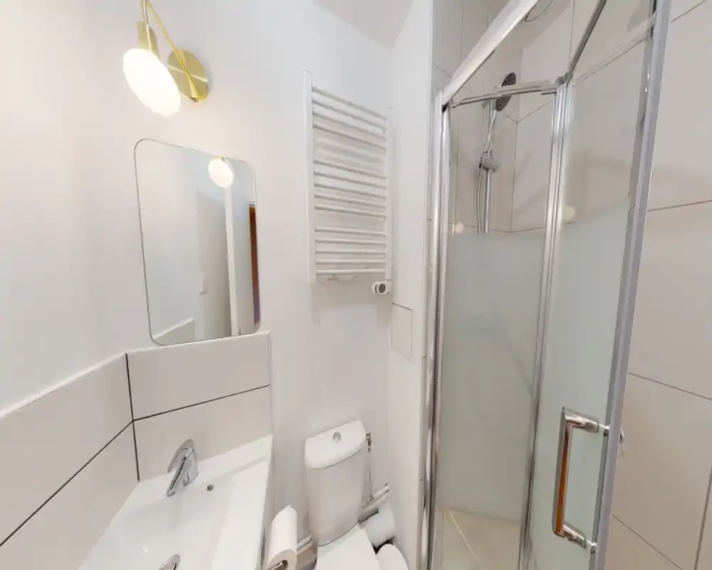 Baño moderno y compacto con ducha de esquina de vidrio, radiador toallero mural, lavabo y inodoro en acabado blanco y limpio.