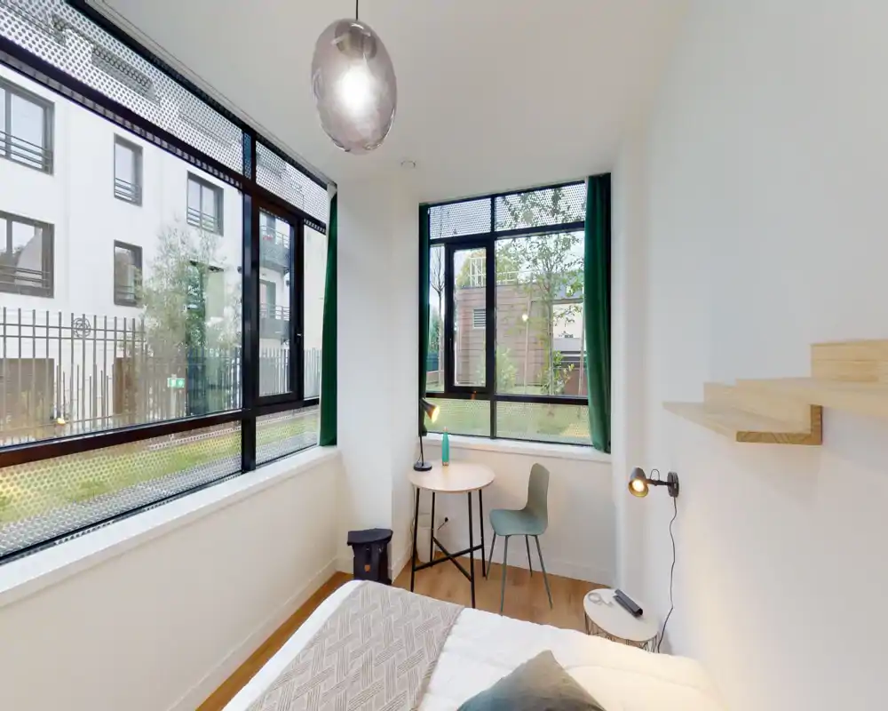 Dormitorio moderno y luminoso con grandes ventanales, cama cómoda con manta decorativa, pequeña mesa redonda y silla junto a la ventana — ideal por su luz natural y vistas a la ciudad.