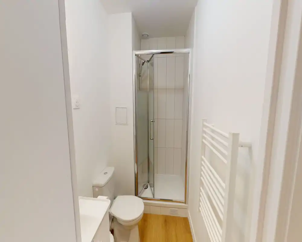 Baño compacto y moderno con cabina de ducha en esquina, inodoro y toallero calefactado; azulejos blancos y suelo efecto madera para un aspecto limpio y minimalista.