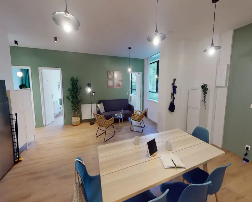 Amplia sala moderna con pared verde, sofá y butacas de mimbre alrededor de una mesa baja. Gran ventana y lámparas colgantes aportan una iluminación clara y acogedora, ideal para relajarse o reuniones informales.