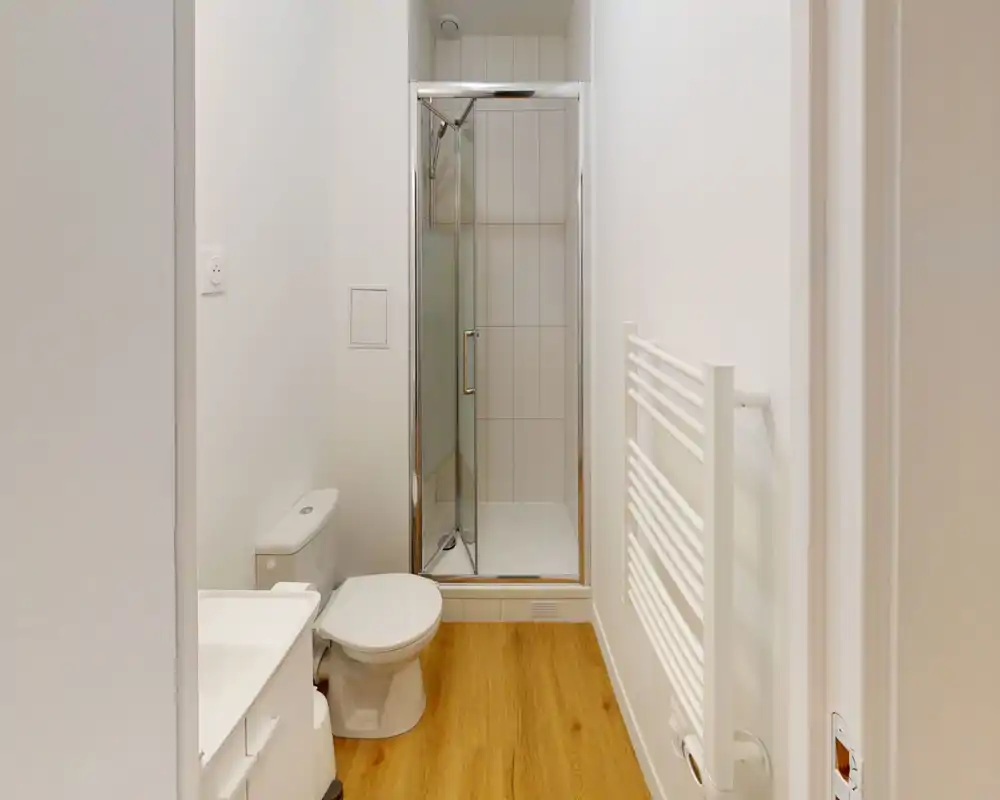 Baño compacto y moderno con cabina de ducha en esquina, inodoro y toallero calefactado sobre suelo efecto madera; decoración luminosa y limpia.