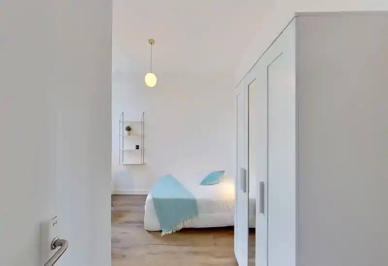 Dormitorio luminoso y minimalista con cama baja con ropa de cama blanca y una manta azul claro; armario con puertas con espejo y estantería de pared pequeña.