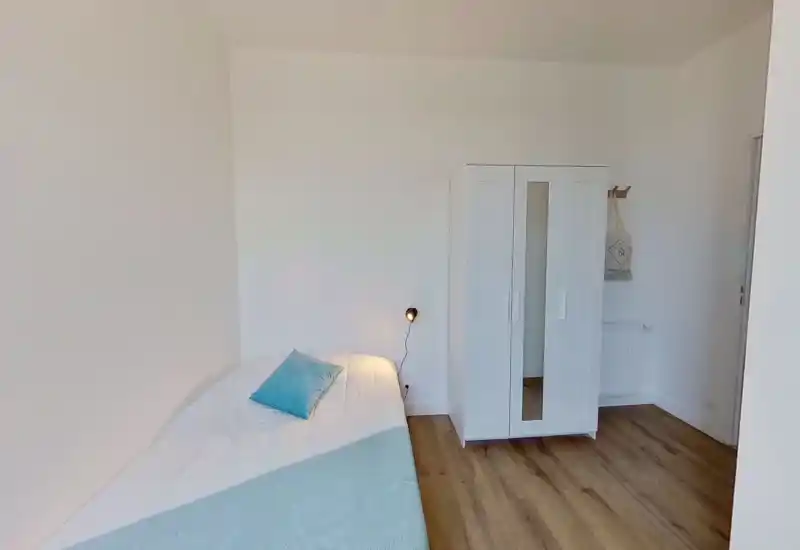 Dormitorio minimalista y luminoso con una cama grande vestida con ropa blanca y una manta azul clara; armario blanco y suelo de madera para un ambiente contemporáneo y tranquilo.