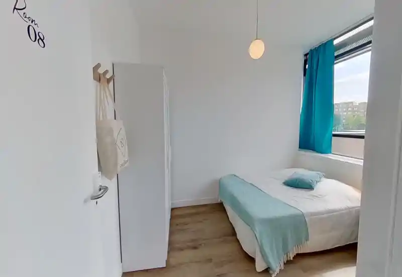 Dormitorio luminoso y minimalista con cama doble vestida en blanco y una manta turquesa; armario y gran ventana que aportan luz natural y espacio de almacenamiento.