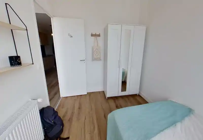 Dormitorio compacto y sencillo con cama individual vestida con una manta turquesa, armario blanco con espejo y suelo de madera. Estilo limpio, luminoso y minimalista, perfecto para alquileres de corta estancia.