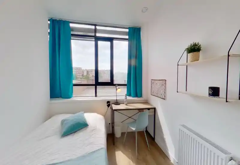 Dormitorio compacto y luminoso con cama individual junto a una gran ventana, cortinas turquesa y un pequeño escritorio — ideal para estudiantes o estancias cortas.