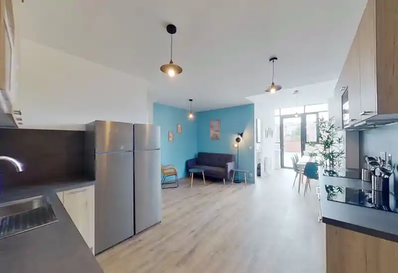 Amplia sala de estar de concepto abierto integrada con la cocina. La habitación incluye un sofá cómodo frente a una pared azul, lámparas colgantes modernas y grandes ventanales que aportan mucha luz natural.