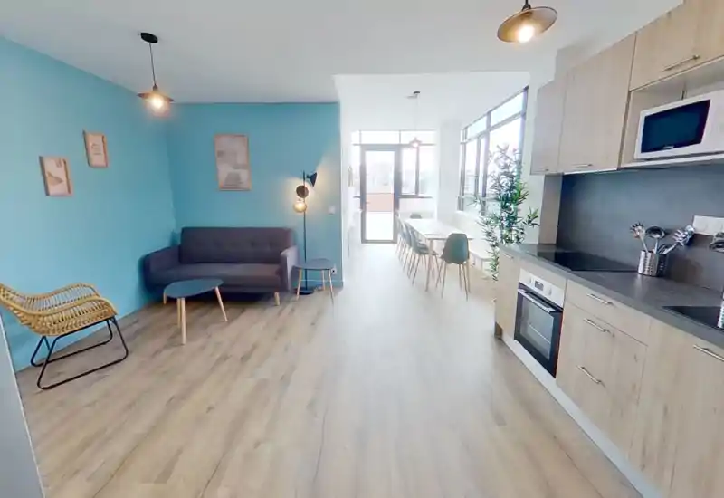 Espacio de salón de planta abierta, luminoso y moderno, con sofá gris, sillón de mimbre, mesas auxiliares y cocina/comedor integrada — perfecto para socializar y relajarse.