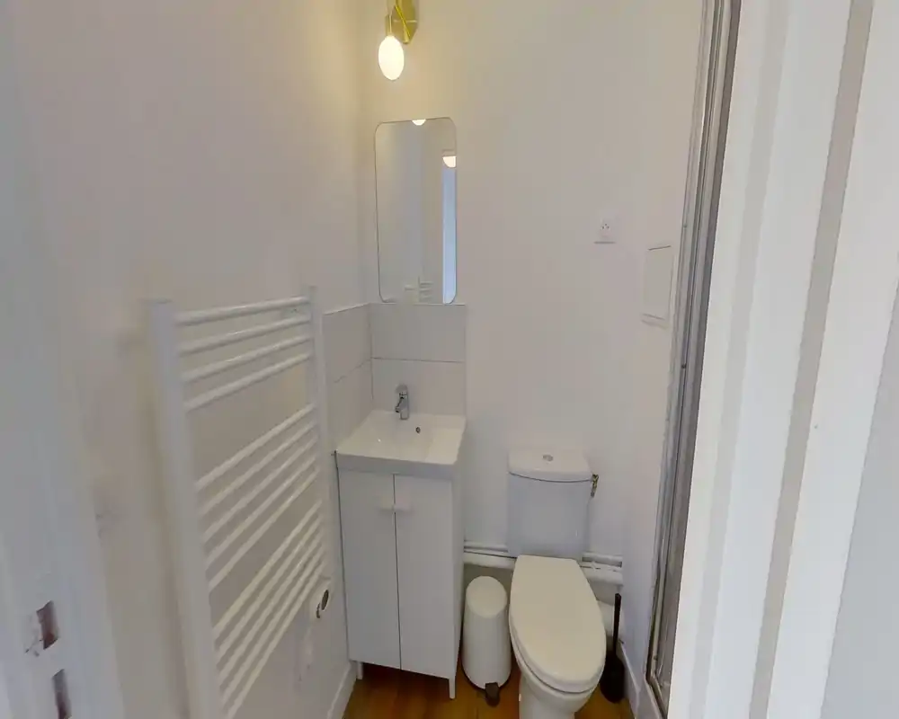 Bagno compatto e luminoso con mobile lavabo, specchio, WC e scaldasalviette — finiture bianche pulite e pavimento effetto legno.