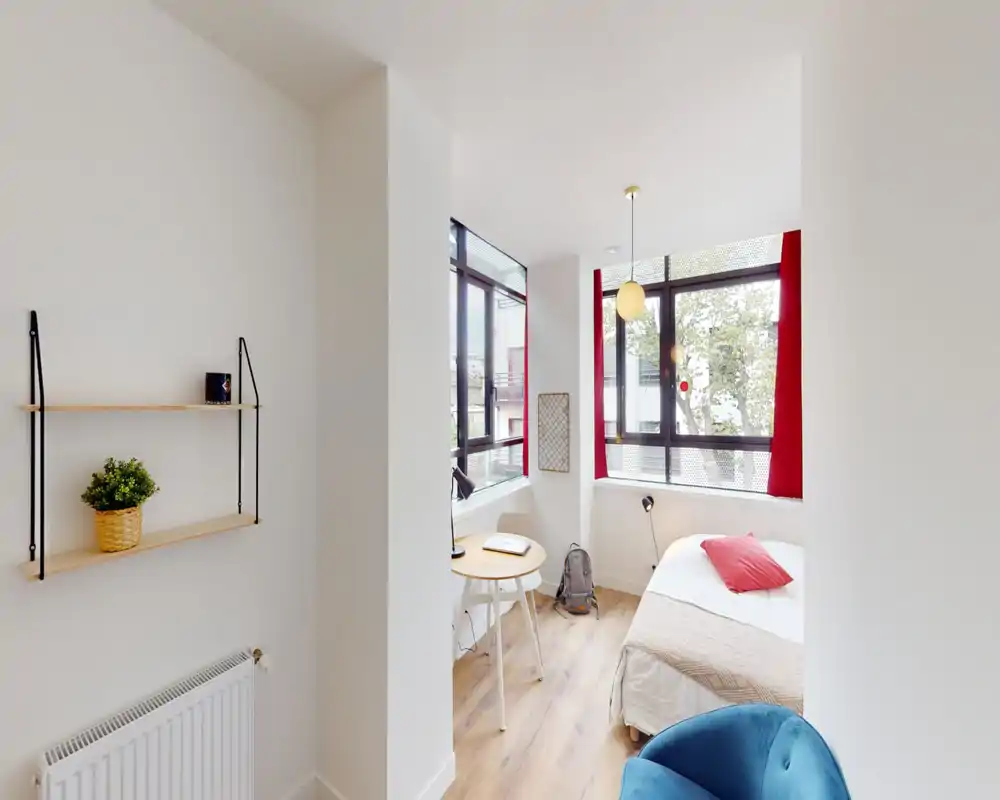 Chambre moderne et lumineuse avec lit bien fait près de grandes fenêtres, petite table de travail et parquet chaleureux — parfaite pour un studio cosy.