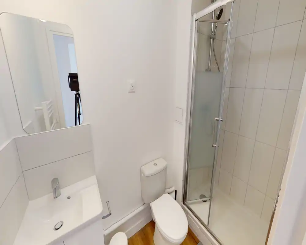 Salle de bain moderne et compacte avec douche carrelée, WC et lavabo. Finitions blanches nettes et porte de douche en verre dépoli.