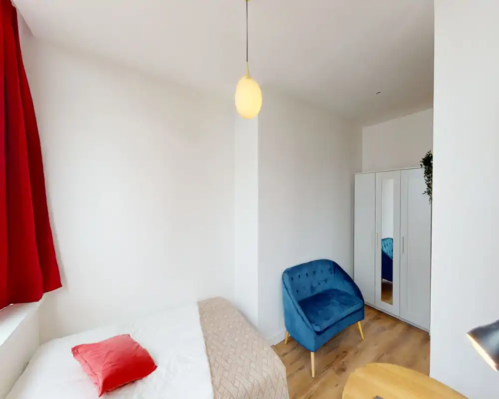 Chambre lumineuse et minimaliste avec lit blanc, accents rouges et un petit canapé bleu confortable ; parquet clair et armoire à miroir pour le rangement et la lumière.