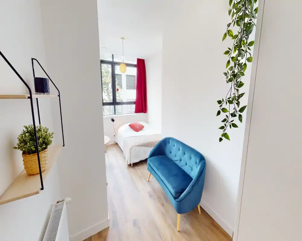 Chambre simple moderne et lumineuse avec lit bien fait près d’une grande fenêtre, sol en bois clair et un petit canapé bleu apportant un coin salon chaleureux.