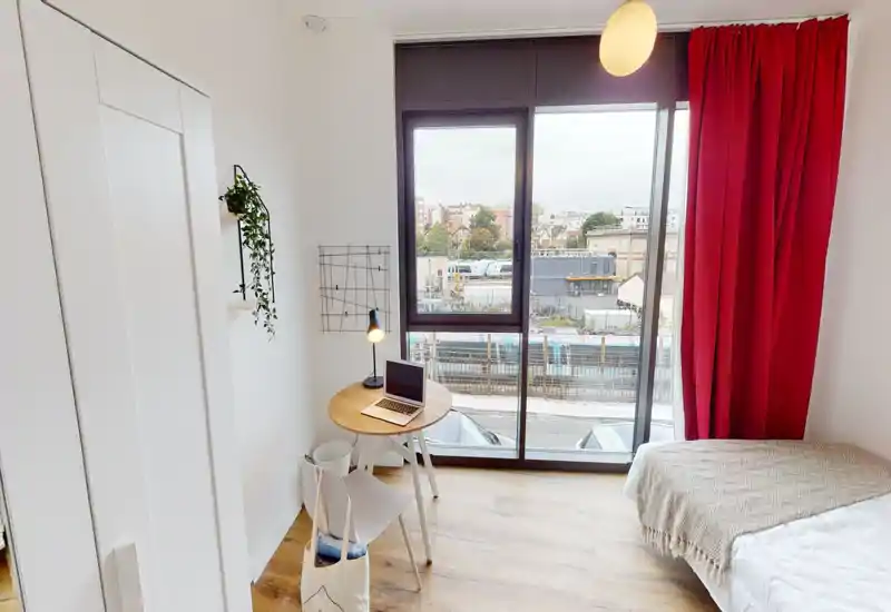 Dormitorio pequeño y luminoso con cama individual junto a una gran ventana de suelo a techo, una mesa redonda compacta con lámpara y portátil, y cortinas rojas que aportan un acento cálido.